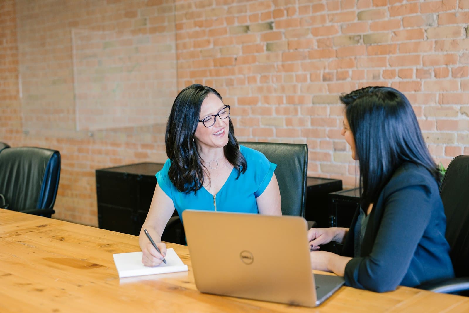 Two women talking across a table, one taking notes beside a laptop
